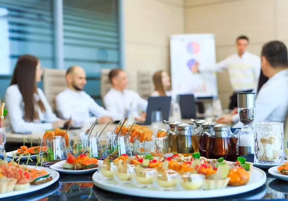 A table of food and drink for an office meeting taking place in the background.