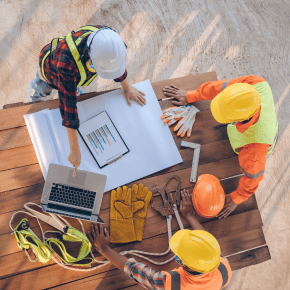 Construction workers wearing hard hats and high-vis vests gathered around a table and looking at a laptop.