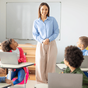 A teacher stands to face children at their desks with laptops.