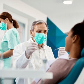 A dentist wearing a face mask holds tools and his patient sits in the chair.