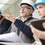 People wearing hard hats and holding a project blueprint. The man in the middle points to something out of frame.